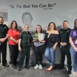 A group of people stands in front of a wall with a quote, "Be The Best You Can Be!" by Buddy Taylor They include uniformed officers and civilians One person in the center holds a plaque and smiles