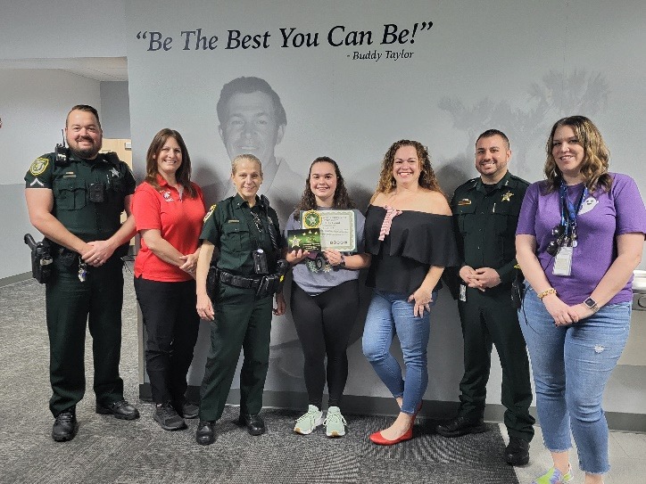 A group of people stands in front of a wall with a quote, "Be The Best You Can Be!" by Buddy Taylor They include uniformed officers and civilians One person in the center holds a plaque and smiles