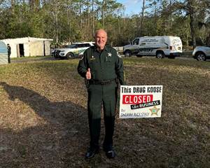 A law enforcement officer wearing a green uniform stands on grass, smiling and giving a thumbs up Next to him is a sign that reads, "This drug house closed for business " Two police vehicles are parked in the background