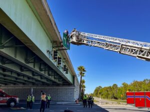 Emergency responders using a fire truck ladder to rescue a person from the edge of a highway overpass Several officials are gathered below, and emergency vehicles are parked nearby Clear blue sky in the background