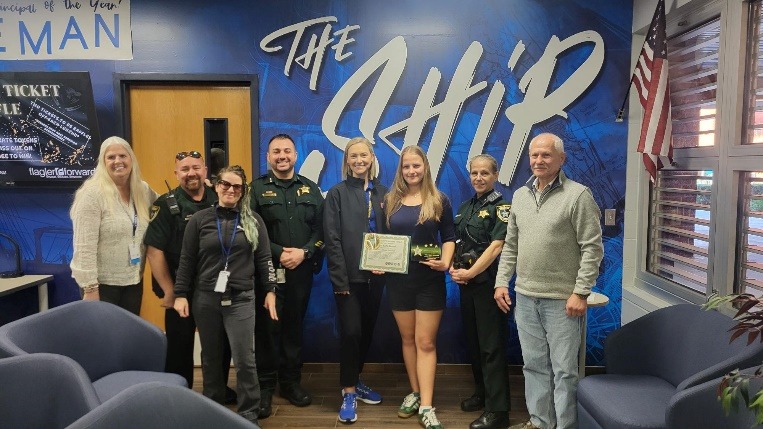 A group of people poses indoors in front of a large sign that says "The Ship " One person holds a certificate The group includes individuals in uniform and casual clothing An American flag is visible in the corner