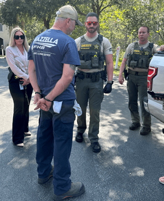 A man in a cap, blue shirt, and jeans is talking to four uniformed officers wearing tactical vests outdoors A vehicle is partially visible on the right, and trees are in the background