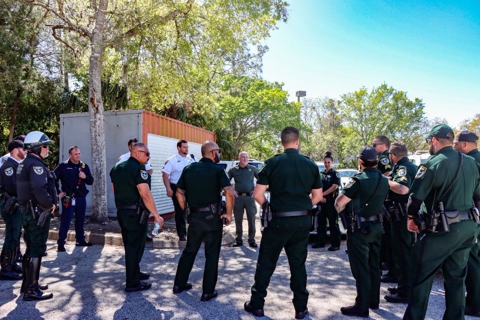 A group of police officers in uniform stand in a circle outdoors, listening attentively to a person addressing them Trees and a storage container are in the background, and it is a sunny day